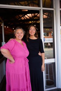 A woman wearing pink and a woman wearing black stand at the entrance to a shop front. They are smiling.