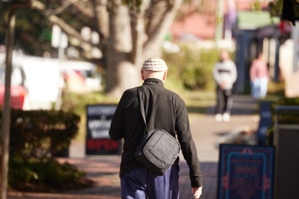 A man with blue trousers and a beanie is walking along the footpath which has a shade awning over the footpath and in front of shops.